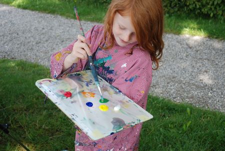 Kleines Mädchen mit roten langen Haaren steht mit einer Farbpalette und Pinsel im Grünen