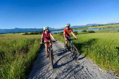 Zwei Radfahrer radeln auf einem Schotterweg umgeben von Wiesen.