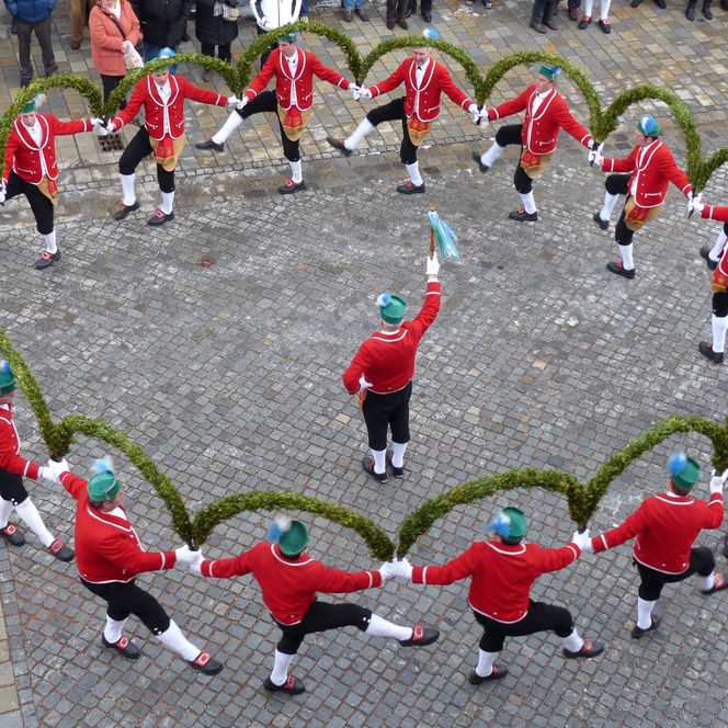 Luftaufnahme des traditionellen Schäfflertanzes mit Schäfflern in roter Tracht und grün geschmückten Bögen.