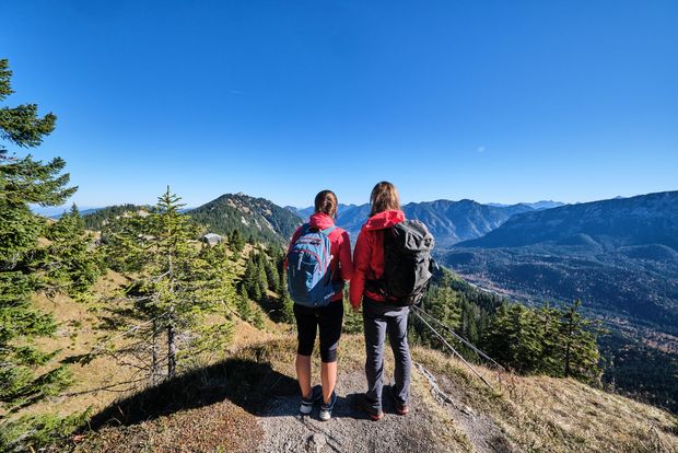 Zwei Personen mit Wanderausrüstung blicken von einem Aussichtspunkt am Wanderweg in die Berge der Zugspitz Region. Die Personen sieht man von hinten.