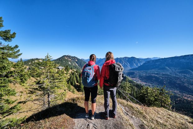 Zwei Personen mit Wanderausrüstung blicken von einem Aussichtspunkt am Wanderweg in die Berge der Zugspitz Region. Die Personen sieht man von hintn.
